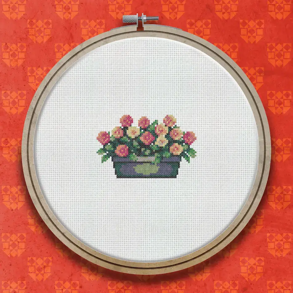 Cross stitch design of pink roses with green foliage growing in a dark rectangular garden trough, shown against a white background.