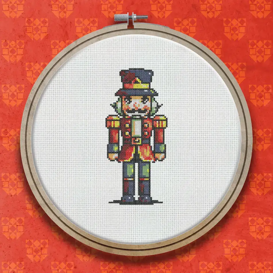 Cross stitch nutcracker soldier wearing a tall red and gold decorated hat, red coat with gold trim, blue trousers, and black boots, standing upright facing forward.