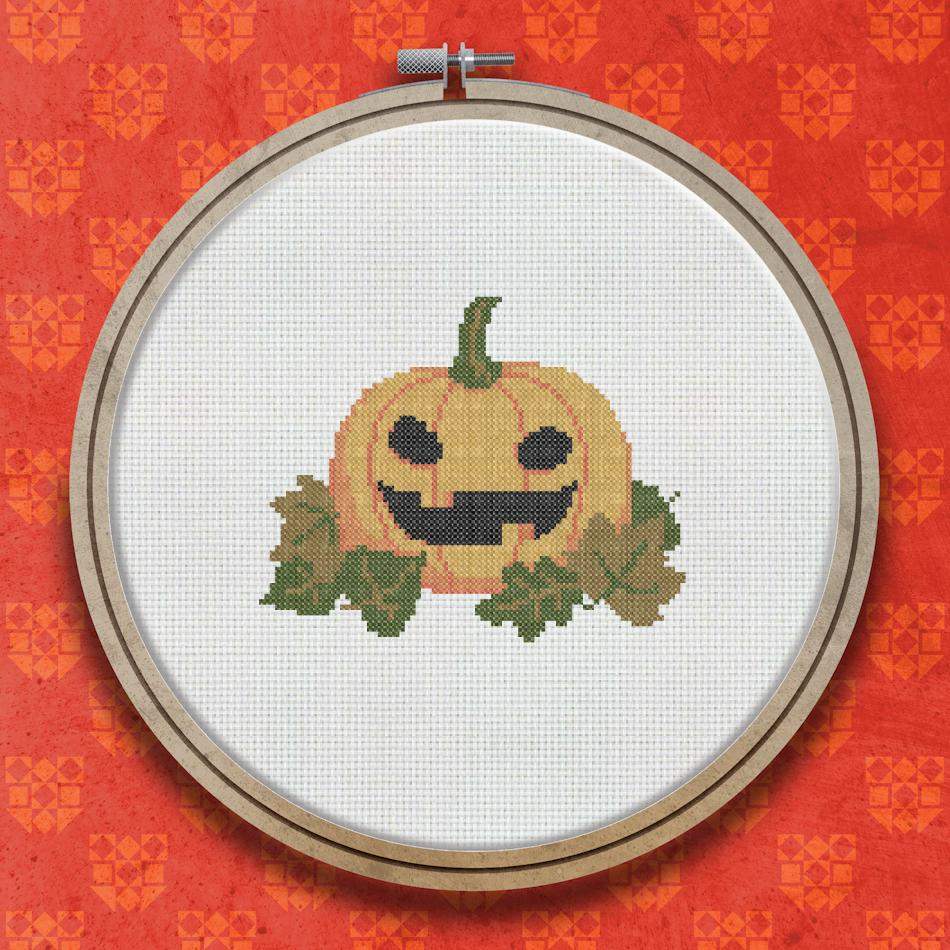 Cross stitch design of a smiling orange jack-o-lantern pumpkin with a carved grinning face, surrounded by green leaves.