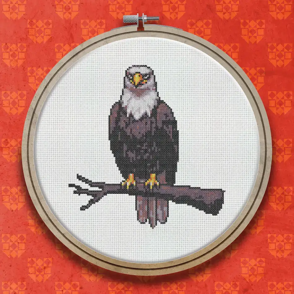 Cross stitch pattern of a bald eagle with a white head and dark brown body perched on a branch, facing forward.