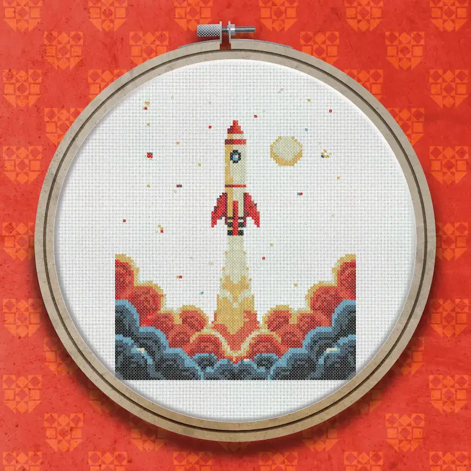 Cross stitch design of a red and white retro rocket launching upward with a large orange and red flame plume below, a small golden moon in the upper right corner.