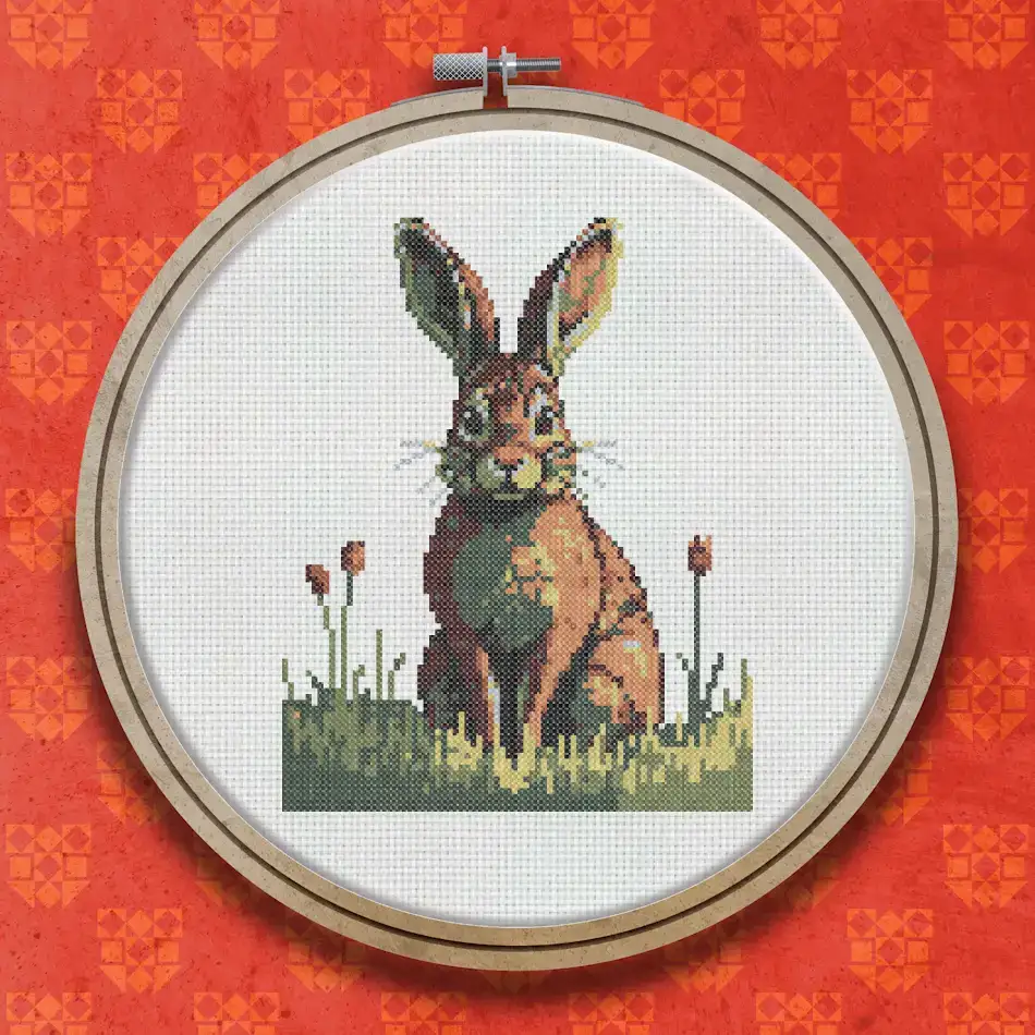 Cross stitch hare sitting upright in green grass with small red wildflowers, rendered in warm browns, tans and earthy tones