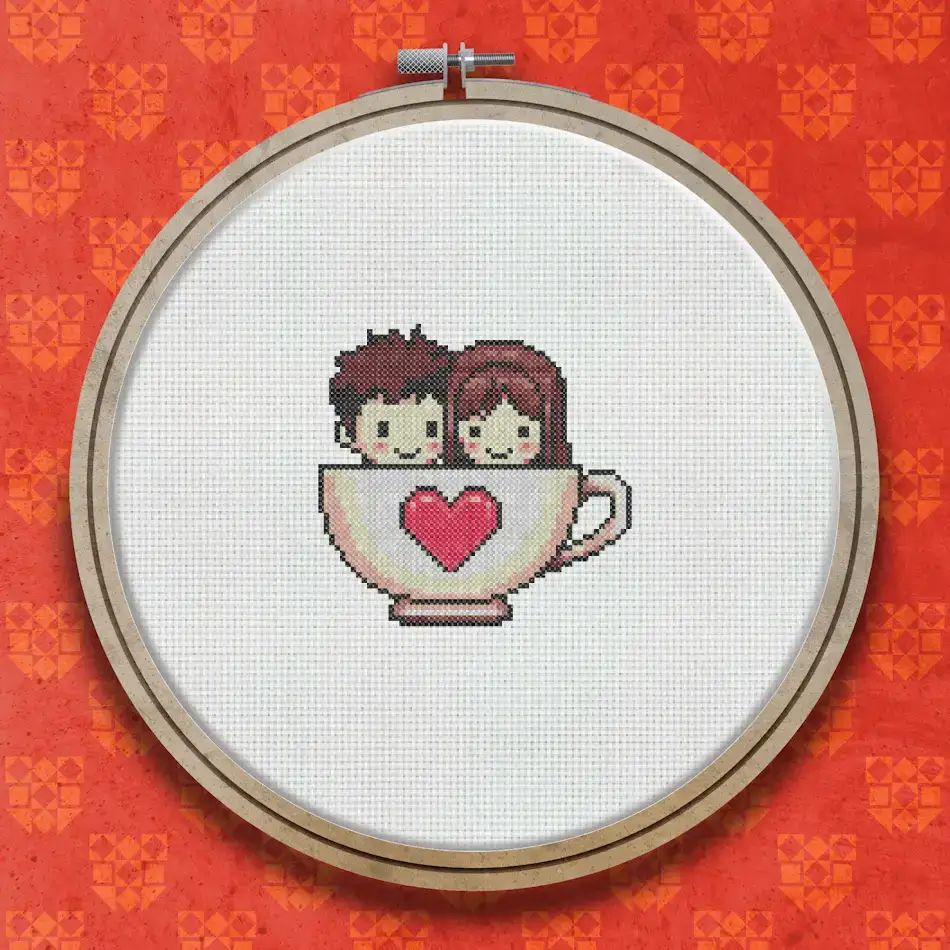 Cross stitch design of a smiling boy and girl sitting together inside a large white coffee cup decorated with a red heart.