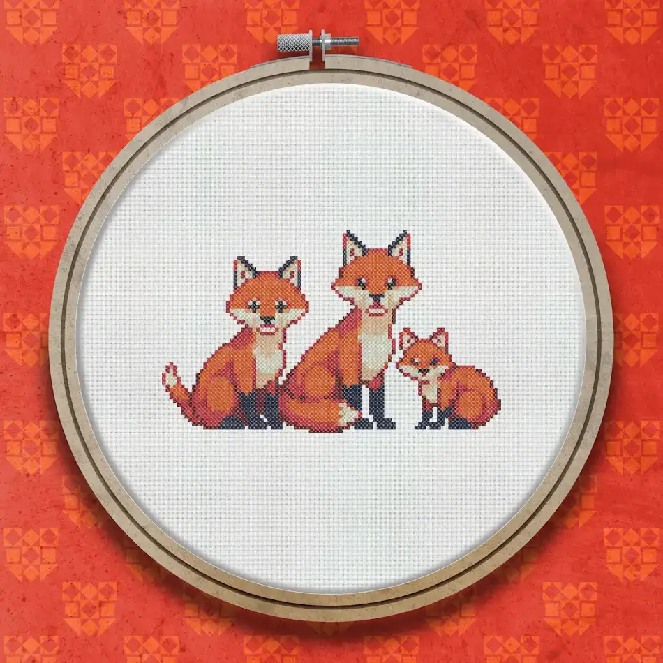 Cross stitch design of two adult foxes and a small fox cub sitting together, stitched in shades of orange, russet, white and black.