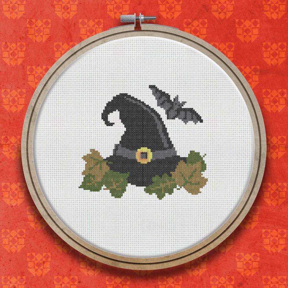 Cross stitch design of a black pointed witches hat with a yellow buckle, a small black bat flying nearby, and green and orange leaves arranged below