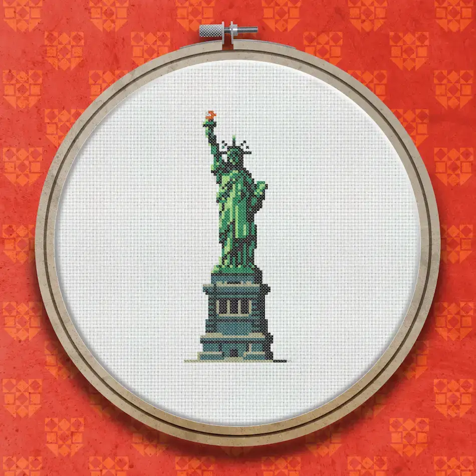 Cross stitch design of the Statue of Liberty in green and grey tones, showing the full figure with torch raised, standing on a layered pedestal base.