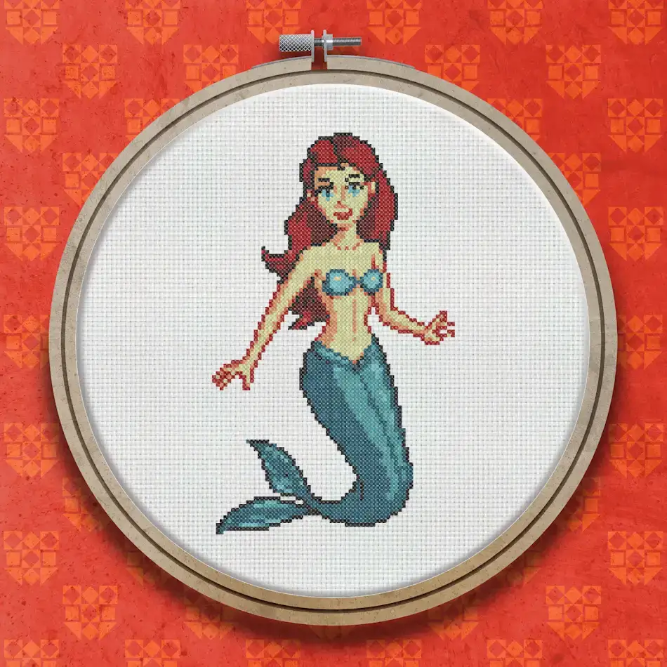 Cross stitch design of a red-haired mermaid with a teal tail and blue-green bikini top, posed upright against a white background.