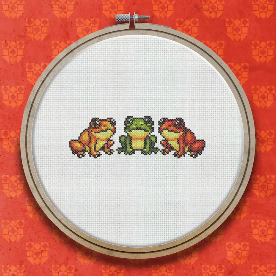 Three small cross stitch frogs in a row, each coloured in shades of orange, green and red with dark outlines, shown on a white background.