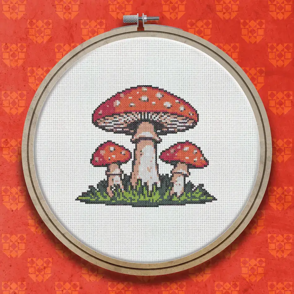Cross stitch pattern showing three red and white spotted toadstools of varying heights clustered together on a patch of green grass