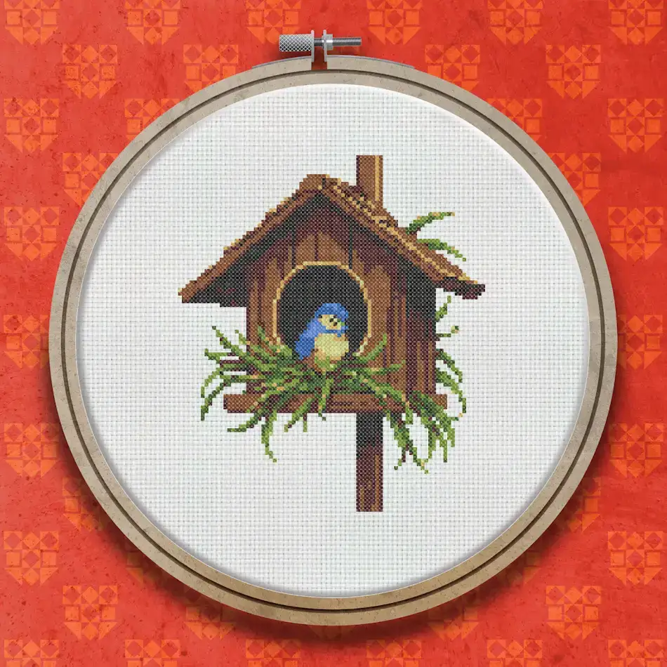 Cross stitch design of a wooden bird nesting box on a post with green leaves, featuring a small blue and orange bird at the entrance hole.