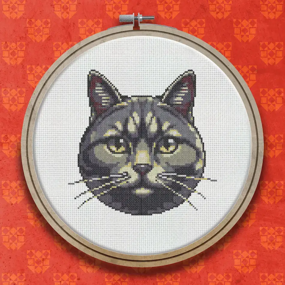 Cross stitch portrait of a grey tabby British Shorthair cat with round amber eyes, bold facial markings and a white muzzle on a dark background.