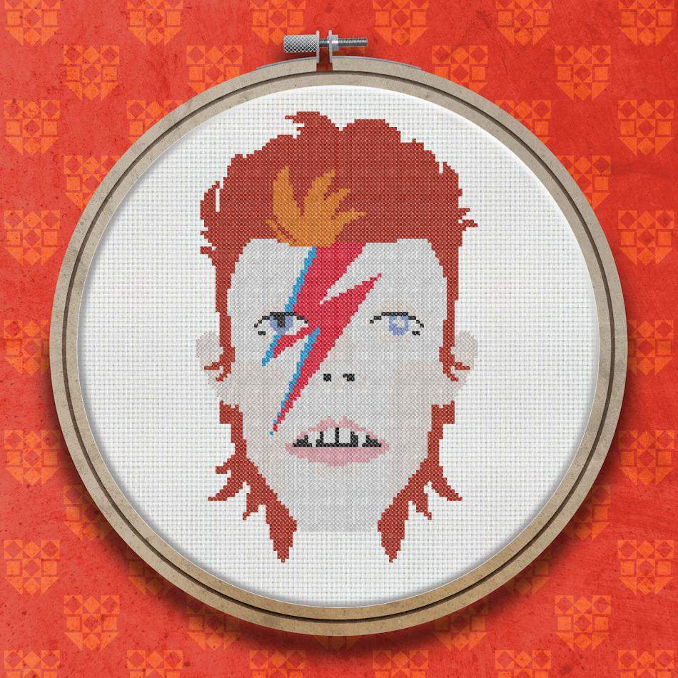 Cross stitch portrait of David Bowie with wild red hair, a red and blue lightning bolt across his face, and pale skin against a white background.