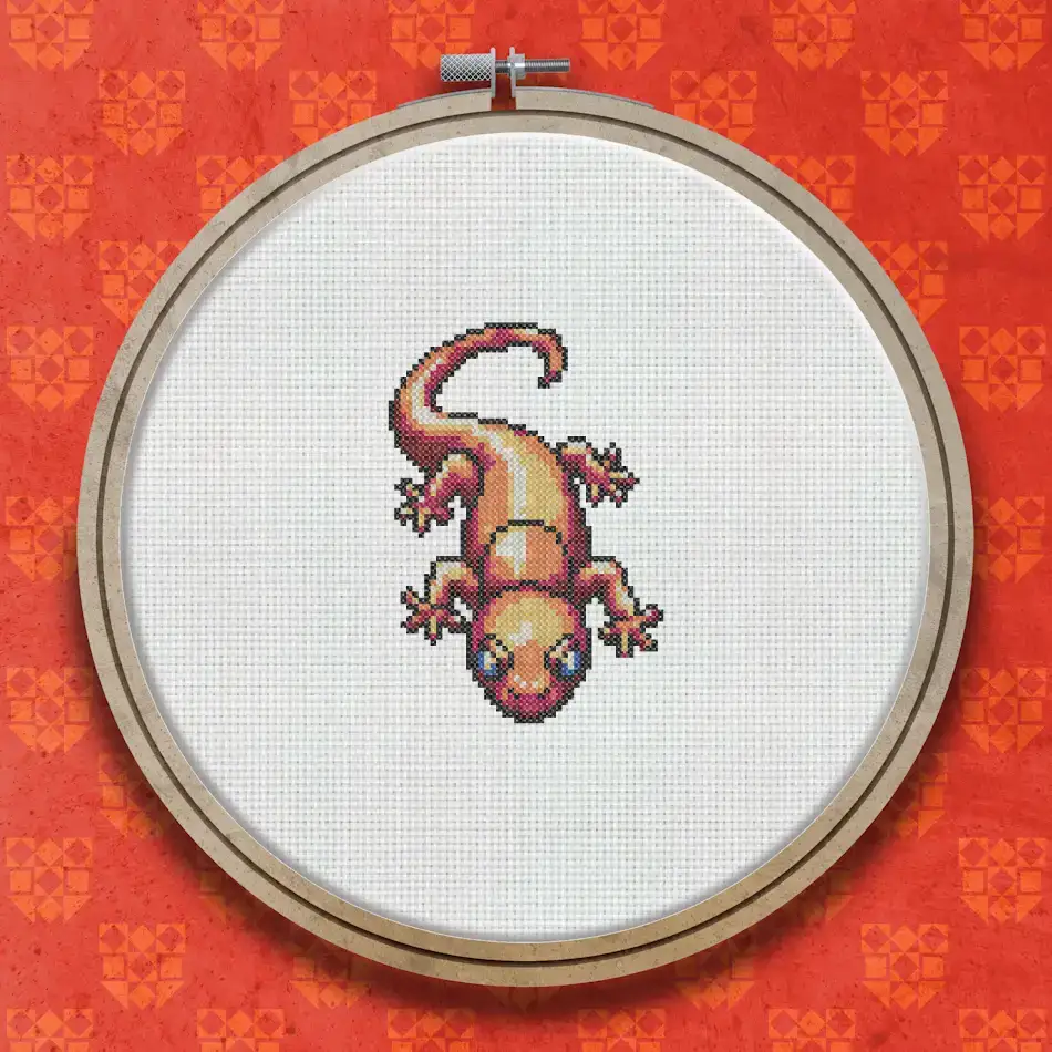 Cross stitch gecko with a vivid red and orange body, darker mottled markings, a long curving tail, and four splayed feet on a white background.