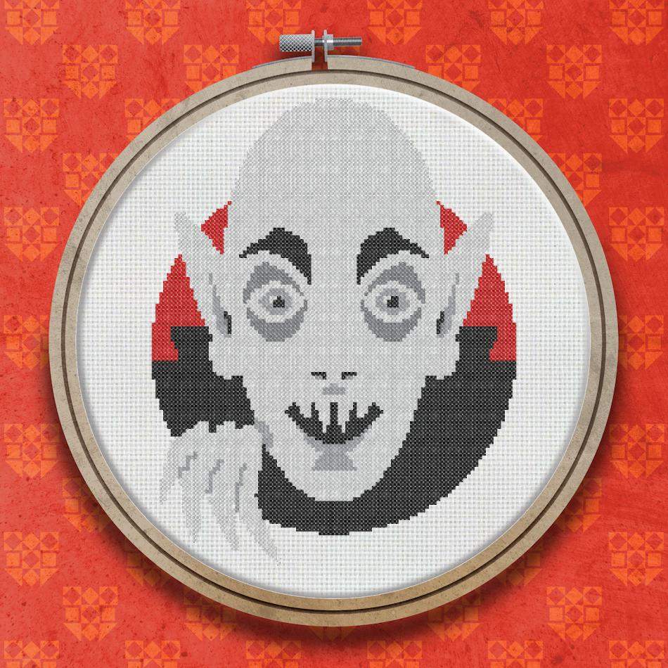 Cross stitch design of a pale vampire face with dark cape, pointed ears, red-lined collar, clawed hand, and an open-mouthed fanged expression.