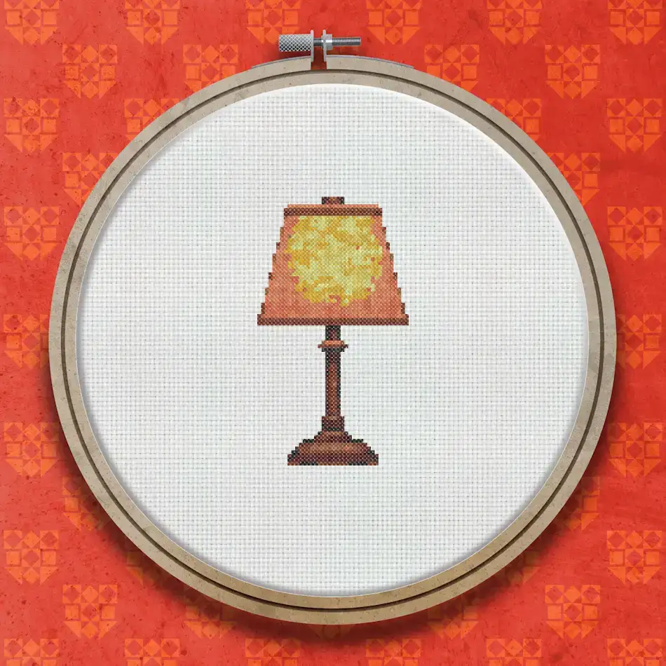 Cross stitch design of a table lamp with a warm amber orange shade and a dark reddish-brown turned base on a plain background.