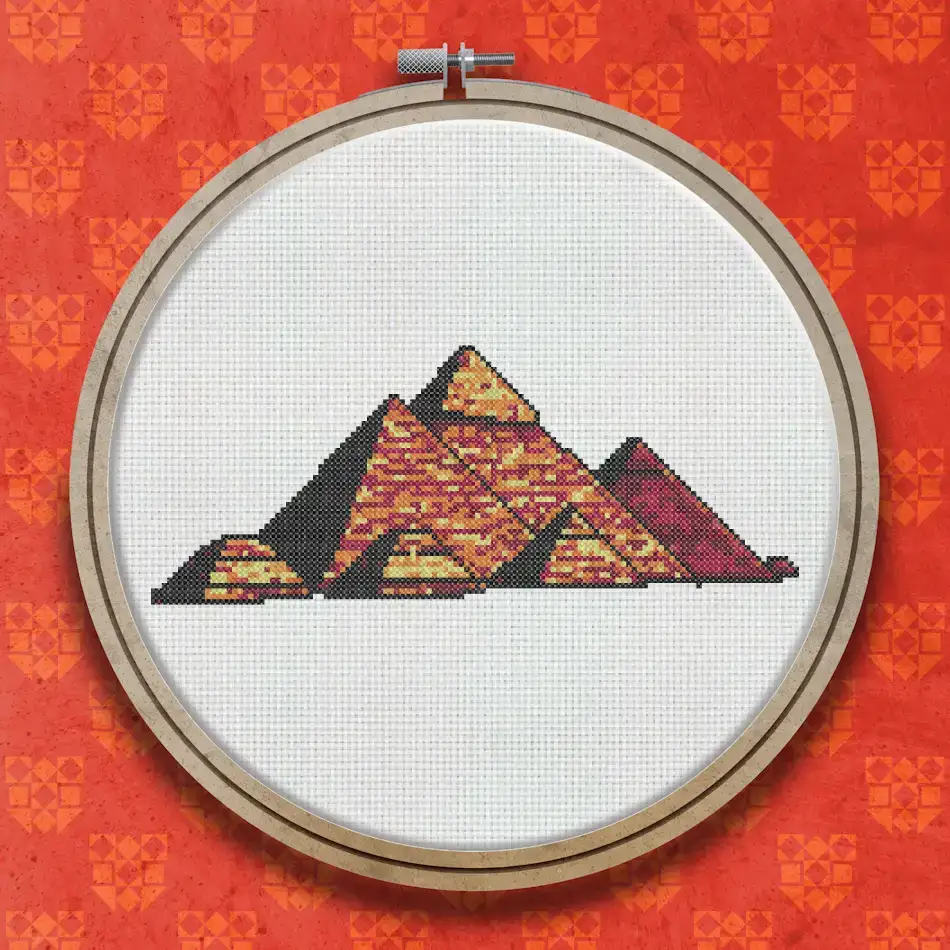 Cross stitch design showing three pyramids in shades of orange, gold and brown against a dark red sky, with a small red pyramid shape to the right.